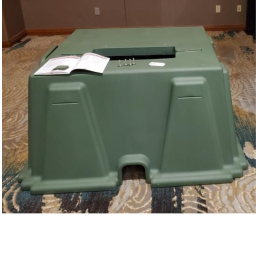 A large green plastic cover stands over a tool on a patterned carpet. An instruction manual and some screws are placed on top of the cover, next to what looks like a Transformer Pad. The background features a brown wall and a wooden baseboard.