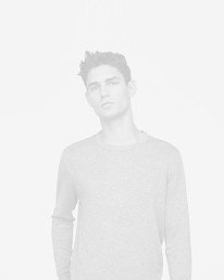 A man with short, dark hair wearing a light gray long-sleeved shirt stands with a serious expression against a white background.