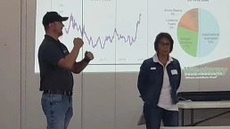 Two people are presenting in front of a projector screen displaying a line graph and a pie chart. The man on the left is gesturing with his hands while speaking, and the woman on the right stands by, listening attentively. They are in a classroom or seminar room.