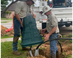 Two utility workers are installing a green plastic enclosure over cables protruding from a hole in the ground, securing electric pedestals. They are wearing helmets and work uniforms. A heavy-duty vehicle with equipment is parked nearby, while trees and a residential area are visible in the background.