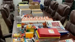 A conference room table filled with supplies including notebooks, glue sticks, markers, colored pencils, scissors, folders, and paper stacks. Leather chairs surround the table. The walls display a large mission statement and logos of various companies.