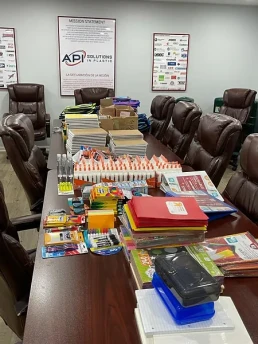 A conference room table filled with supplies including notebooks, glue sticks, markers, colored pencils, scissors, folders, and paper stacks. Leather chairs surround the table. The walls display a large mission statement and logos of various companies.