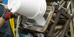 A person wearing red and black gloves is pouring a white substance from a large white bucket into a rectangular industrial machine. The machine, supported by metal framework, appears to be part of manufacturing or production services.