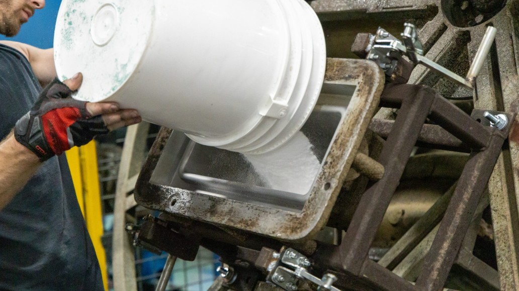 A person wearing red and black gloves carefully pours a white substance from a large white plastic bucket into a metal container that is part of an industrial machine. The machine, about which much can be said, has a rusted and mechanical appearance, complete with various bolts and brackets.
