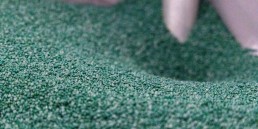 Close-up of green granular fertilizer being poured from a spout into a container. The small granules are uniformly sized, creating a textured surface. The spout is positioned in the upper part of the image, directing the flow of the fertilizer.