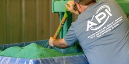 A person wearing a gray T-shirt with lettering is using a tool to mix or adjust a large container filled with green powdered material, likely as part of industrial services in a manufacturing setting.
