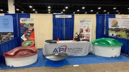 A trade show booth features the company API Solutions in Plastic. The setup includes a table with a banner, brochures, and samples. Surrounding the table are several large plastic structures in red, green, and blue. Two posters display product information.