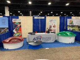 A trade show booth features the company API Solutions in Plastic. The setup includes a table with a banner, brochures, and samples. Surrounding the table are several large plastic structures in red, green, and blue. Two posters display product information.