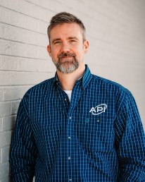A man with short gray hair and a beard is standing against a light gray brick wall. He is wearing a blue checkered shirt with the 