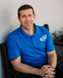 A man sits outside against a white brick wall, wearing a bright blue polo shirt with an 