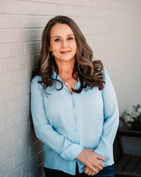 A woman with long, wavy brown hair is standing against a white brick wall, wearing a light blue blouse and blue jeans. She is smiling softly and has her hands clasped in front. She looks like she could be part of the API Team, ready to tackle projects with a calm yet confident demeanor.