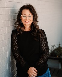 A woman with wavy brown hair and glasses stands against a white brick wall, smiling. She is wearing a black top with patterned sleeves and blue jeans, her hands clasped in front of her. Behind her, there's a blurred plant, adding a touch of green to the scene. She looks like she could be part of an API team photo shoot.