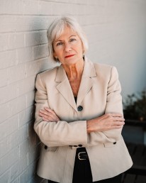 A senior woman with short gray hair and wearing a beige blazer stands with her arms crossed against a white brick wall. She has a serious expression on her face, embodying the resilience of the API Team she leads.