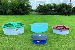 A green field, essential for agriculture, features four different water troughs for livestock placed on the grass. The troughs vary in design and color: a green-topped trough, a light blue open trough, a maroon-topped trough, and a blue trough with a black lid. Trees are visible in the background.