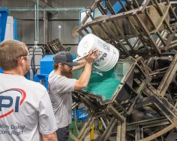 A man with a beard and glasses pours green material from a white bucket into a large piece of industrial equipment. He is observed by another man wearing a white API-branded shirt. The setting appears to be a busy factory or industrial facility.