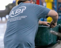 A man in a blue shirt and a gray cap is working in a warehouse or industrial setting. His shirt, emblazoned with a company logo on the back, signifies the reliable services offered by his team. He is handling a large green object on a workbench, with brightly colored shelves and equipment visible in the background.