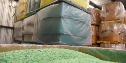 Photograph showing a warehouse with stacked plastic bins covered in clear plastic on pallets. In the foreground, there is a container filled with bright green granulated material, showcasing the efficient storage services. The walls and ceiling structure of the warehouse are visible.