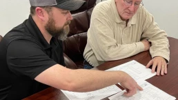 Two men are seated at a wooden table reviewing architectural blueprints. The man on the left, wearing a black polo shirt and gray cap, is pointing at the plans. The older man on the right, wearing glasses and a beige shirt, is attentively looking at the documents.