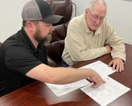 Two men are seated at a wooden table reviewing architectural blueprints. The man on the left, wearing a black polo shirt and gray cap, is pointing at the plans. The older man on the right, wearing glasses and a beige shirt, is attentively looking at the documents.