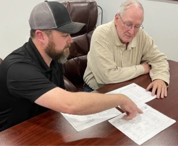 Two men are seated at a wooden table reviewing architectural blueprints. The man on the left, wearing a black polo shirt and gray cap, is pointing at the plans. The older man on the right, wearing glasses and a beige shirt, is attentively looking at the documents.
