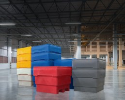 A group of large plastic storage bins in various colors (blue, yellow, red, grey) is stacked in the foreground of a spacious, well-lit industrial warehouse with high ceilings and metal support beams. The shiny floor acts like advanced pedestals, reflecting both the bins and lights.