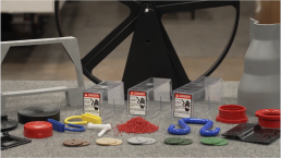 A table displaying various 3D-printed objects of different colors and shapes, including hooks, rings, washers, and blocks. Three transparent boxes with 