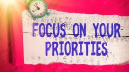 A brightly lit image with a red crumpled paper background features an old-fashioned blue alarm clock and a white wooden sign with large blue text that reads,