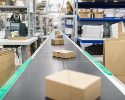 A conveyor belt in a warehouse transports brown rectangular packages, showcasing the efficiency of our logistics services. The surroundings feature shelves stocked with various items, including boxes and packaging supplies. The background is slightly out of focus, highlighting the organized workspace.