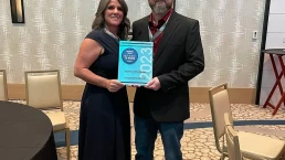 A woman in a blue dress and a man in a black suit stand smiling while holding an award that reads,