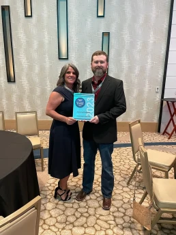 A woman in a blue dress and a man in a black suit stand smiling while holding an award that reads, 