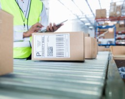 A person in a high-visibility vest uses a tablet while inspecting a brown package labeled 