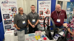 Three people stand behind a table at a trade show booth for API Solutions in Plastic. The table displays plastic products and marketing materials. Two men and one woman wear company-branded shirts and name badges. Booth banners detail the company's molding services.
