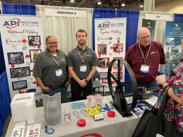 Three people stand behind a table at a trade show booth for API Solutions in Plastic. The table displays plastic products and marketing materials. Two men and one woman wear company-branded shirts and name badges. Booth banners detail the company's molding services.