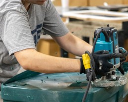 A person wearing safety glasses and gloves operates a power tool to carve or shape a green plastic material in a workshop. The background includes shelves stocked with various tools essential for providing high-quality services.