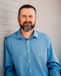 A man with a beard and short hair is standing against a light-colored brick wall. Wearing a blue button-up shirt, he smiles softly at the camera, embodying the approachable spirit of the API Team.